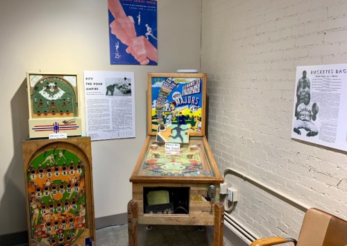 A vintage pinball machine and game displays in a small room, with posters and information on sports history.