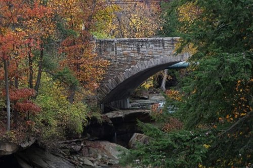 A stone bridge arches over a rocky stream, surrounded by vibrant autumn foliage.