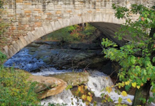 A stone bridge arches over a flowing stream, surrounded by lush greenery and rocky terrain.