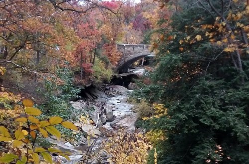 A scenic view of a rocky stream surrounded by vibrant autumn foliage and a stone bridge in the background.