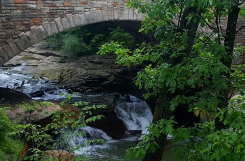 A stone bridge arches over a flowing stream, surrounded by lush green trees and rocky banks.