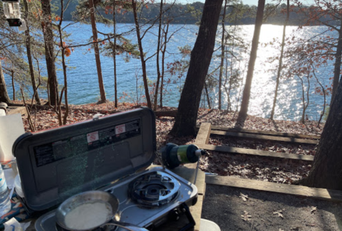 A camping stove with a pot sits on a wooden table, overlooking a serene lake surrounded by trees.