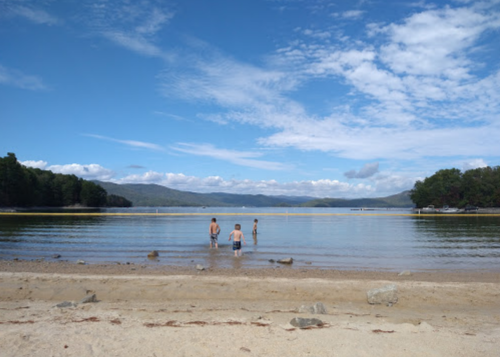 Three people wade in a calm lake on a sunny day, with mountains and a blue sky in the background.