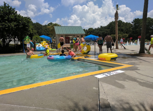 A busy pool area with people enjoying floats, surrounded by trees and blue skies. Sun loungers and umbrellas are visible.