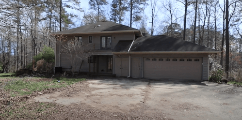 A two-story house surrounded by trees, with a driveway and a garage, set in a wooded area.