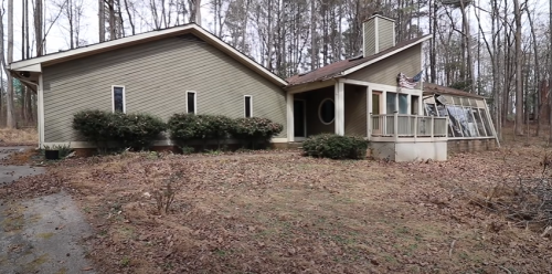 A modern, single-story house surrounded by trees and dry leaves, featuring large windows and a sloped roof.