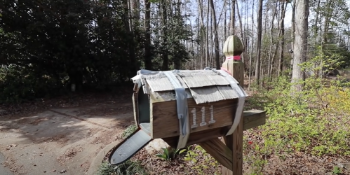 A wooden mailbox with a makeshift cover, surrounded by trees and a gravel path.