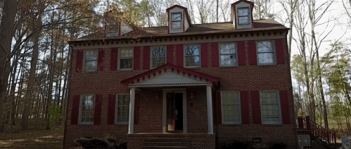 A large brick house with red shutters and a front porch, surrounded by trees in a wooded area.