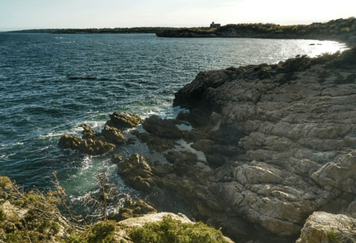 Rocky coastline with waves gently crashing against the shore, under a clear sky and distant land in the background.