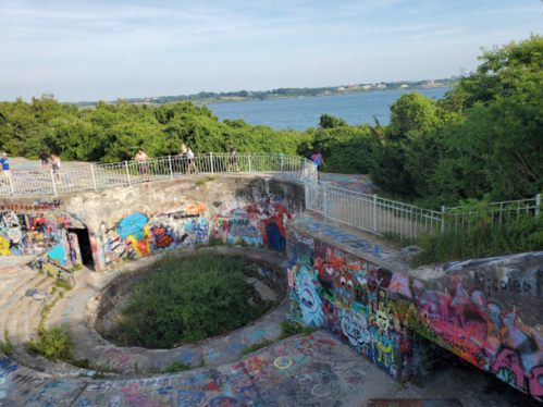 Graffiti-covered ruins overlook a lush landscape and water, with people exploring the area.