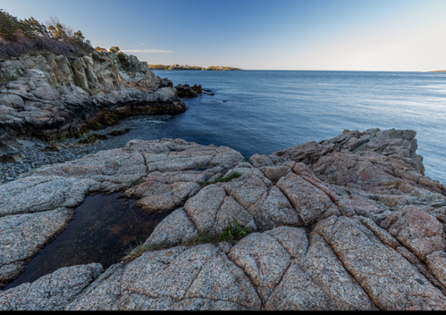 Rocky shoreline with calm blue water and a clear sky, featuring a small tide pool among the stones.
