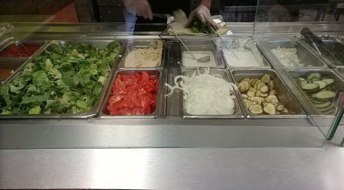 A salad bar with various fresh ingredients: lettuce, tomatoes, onions, cucumbers, and dressings in metal containers.