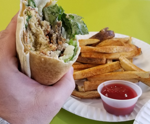 A hand holding a wrapped sandwich with lettuce, next to a plate of golden fries and a small cup of ketchup.