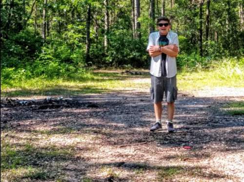 A person stands in a wooded area, wearing sunglasses and a t-shirt, with arms crossed and a path behind them.
