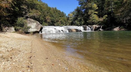 A serene waterfall cascades into a clear pool, surrounded by lush greenery and a sandy shore under a blue sky.