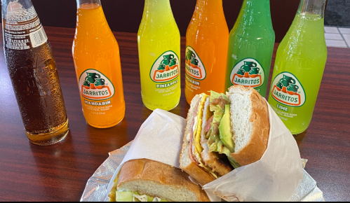 A sandwich with layers of ingredients, surrounded by colorful Jarritos soda bottles in various flavors.