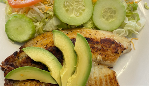 Grilled fish fillet topped with avocado slices, served with cucumber and lettuce salad, and garnished with tomato.