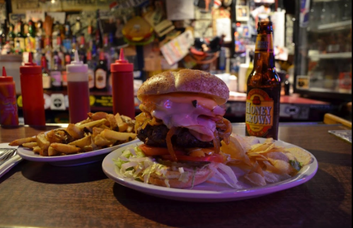 A large burger with toppings, served with fries and a bottle of Honey Brown beer in a lively bar setting.