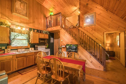 Cozy wooden cabin interior featuring a kitchen, dining area, and staircase with warm lighting and rustic decor.