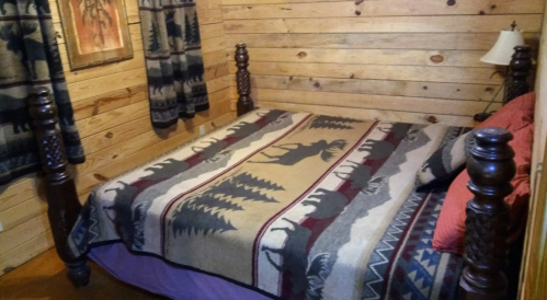 Cozy bedroom with a wooden bed, featuring a patterned blanket and rustic wooden walls. Soft lighting from a lamp.