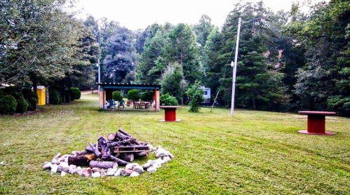A grassy area with a fire pit, surrounded by trees, featuring a gazebo and seating in the background.
