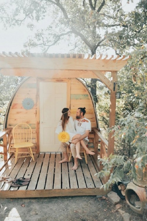 A couple sits on the porch of a cozy wooden cabin, smiling at each other amidst a sunny, natural setting.