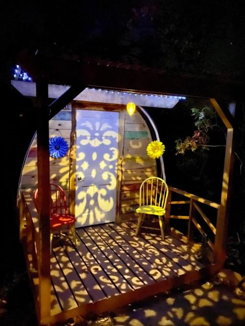 A cozy cabin at night, illuminated with colorful lights and shadows, featuring two chairs on a wooden porch.