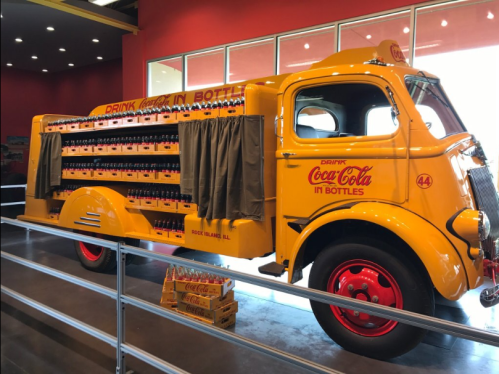 A vintage yellow Coca-Cola delivery truck with bottles displayed on the side, set in a bright indoor space.