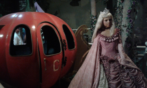 A princess doll in a pink gown stands beside a large pumpkin carriage, set in a whimsical, fairy-tale environment.