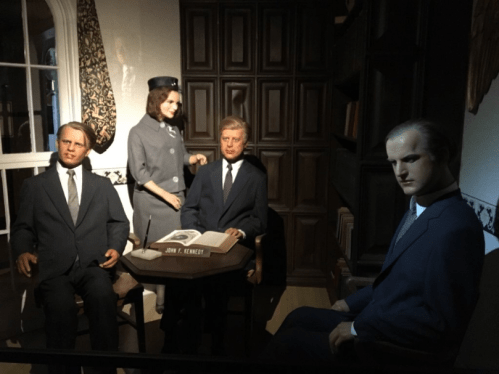 A group of lifelike wax figures seated around a table in a dimly lit room, resembling historical figures.