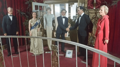 A display of lifelike wax figures of historical figures in formal attire, set against a red curtain backdrop.