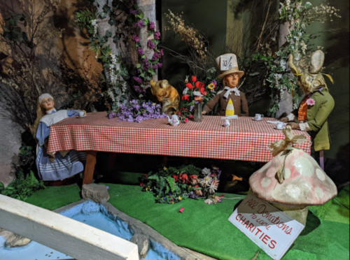 A whimsical tea party scene with dolls, a checkered tablecloth, flowers, and a sign for charity donations.