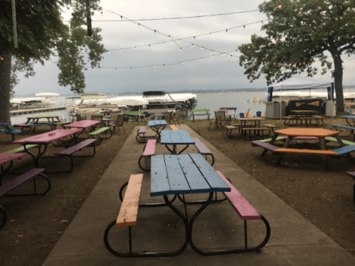Colorful picnic tables line a path by the lake, with boats docked and string lights overhead on a cloudy day.