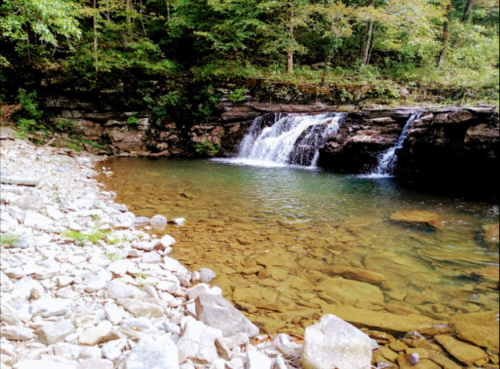 A serene scene of a small waterfall cascading into a clear pool, surrounded by rocky terrain and lush greenery.