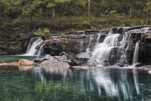 A serene waterfall cascades over rocky ledges into a tranquil pool, surrounded by lush greenery.