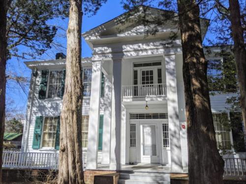 Historic white mansion with green shutters, surrounded by tall trees and a clear blue sky.