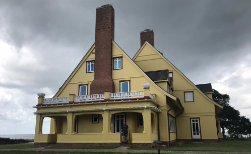 A large yellow house with multiple chimneys and a porch, set against a cloudy sky. A person stands at the entrance.