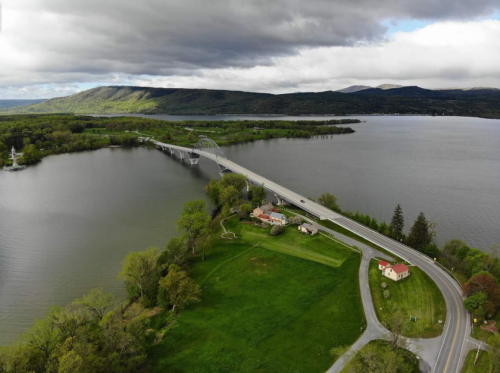 Aerial view of a bridge over a river, surrounded by green fields and hills under a cloudy sky.