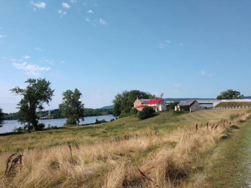 A scenic view of a riverbank with a house and barn, surrounded by tall grass and trees under a clear blue sky.