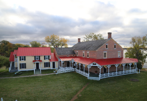 Historic building with a red roof, surrounded by green grass and trees, near a body of water under a cloudy sky.