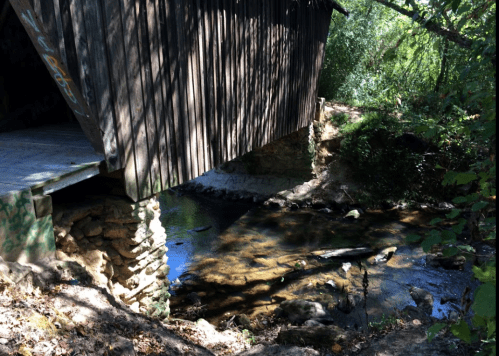 A wooden structure overhangs a small, clear stream surrounded by trees and foliage.