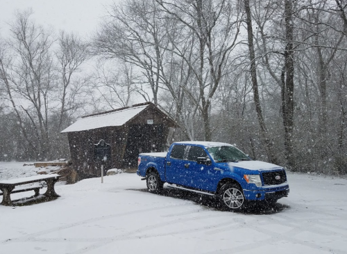 A blue pickup truck parked in a snowy landscape near a small wooden cabin, with snow falling heavily.