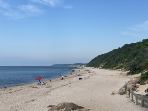 A serene beach scene with a sandy shore, gentle waves, and a few people relaxing under umbrellas. Lush green hills in the background.