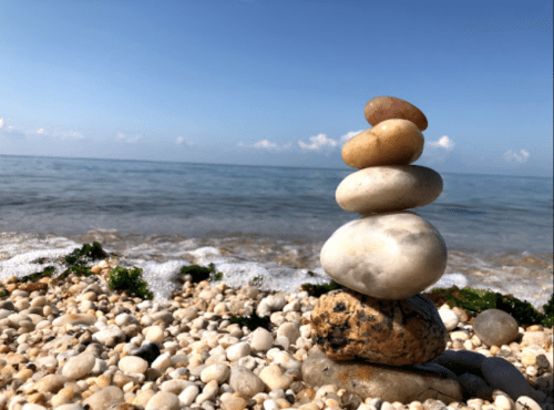 A stack of smooth stones balanced on a beach, with gentle waves and a clear blue sky in the background.