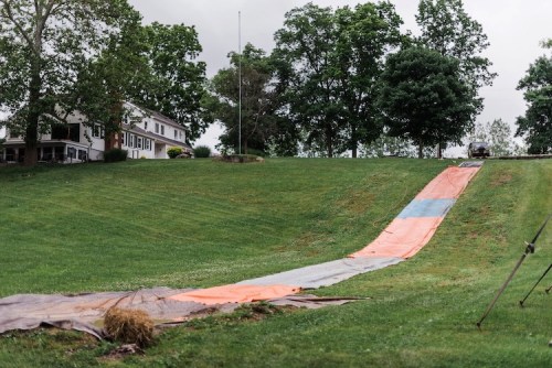 A large, colorful slip-and-slide stretches down a grassy hill, with trees and a house in the background.