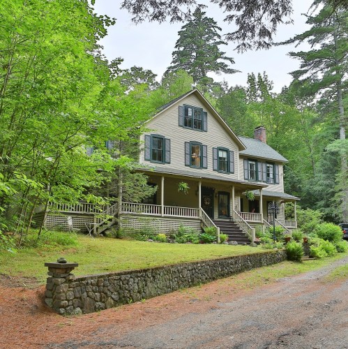 A charming two-story house surrounded by lush greenery and tall trees, with a stone pathway leading to the entrance.