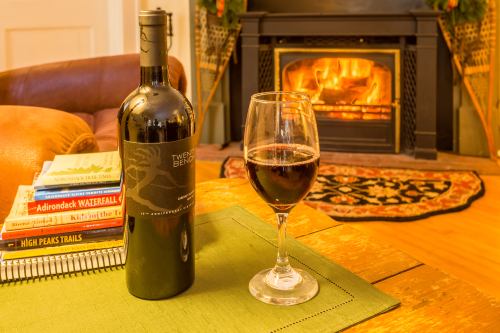 A bottle of red wine and a glass sit on a table near a cozy fireplace, surrounded by books and a warm atmosphere.