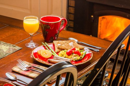 A cozy breakfast setting with toast, eggs, sausage, asparagus, orange, and drinks by a warm fireplace.