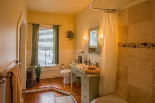 A cozy bathroom with a shower, toilet, sink, and natural light from a window, featuring warm yellow walls and green accents.