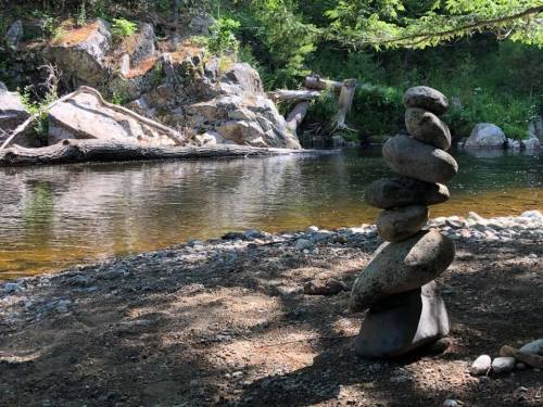 A stack of smooth stones balanced on a riverbank, surrounded by trees and rocky terrain.